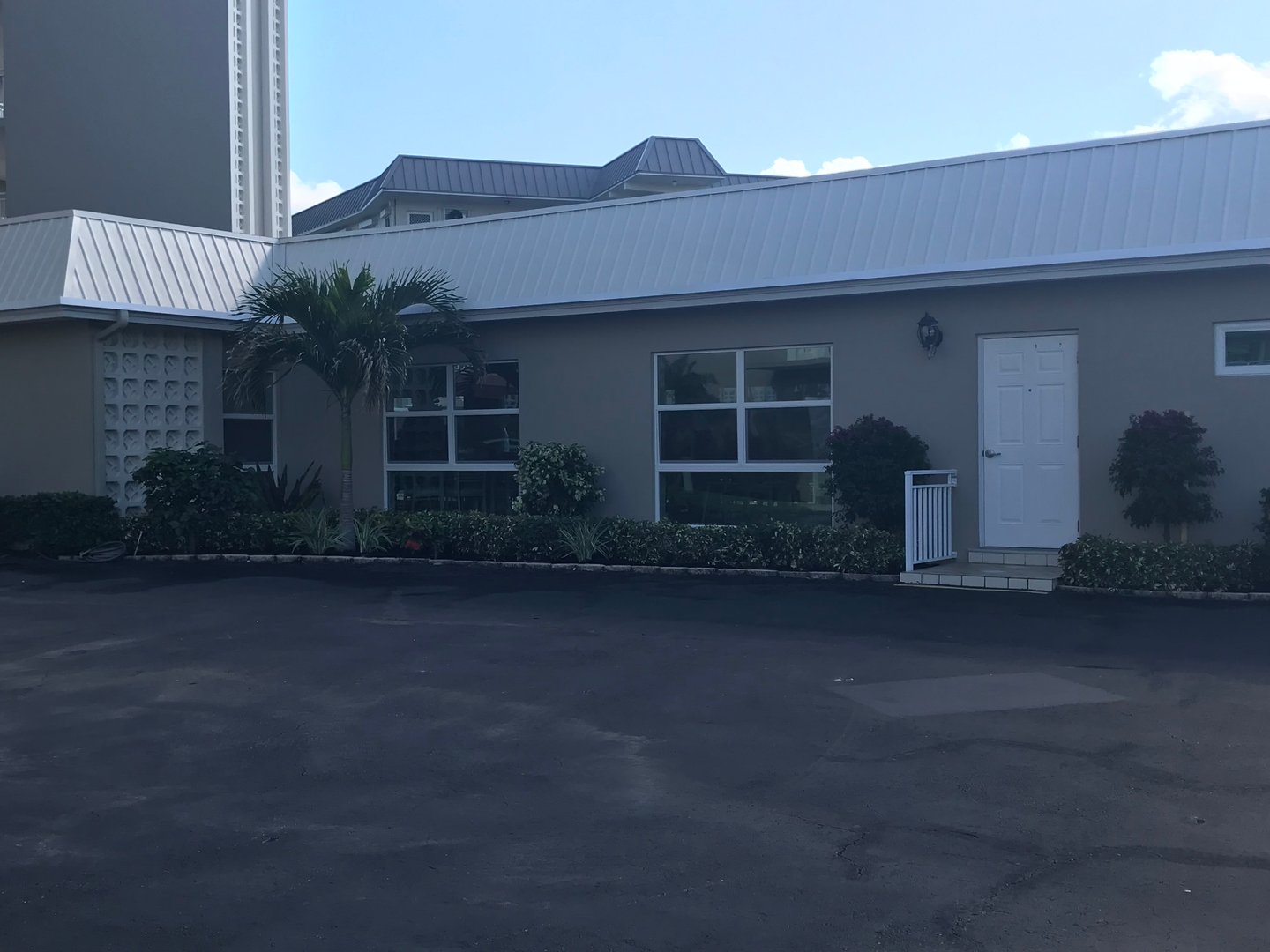 Single-story building with large windows, a white door, and a palm tree in front, set against a clear blue sky.