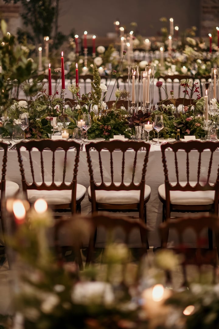 Beautifully decorated wedding reception hall featuring long banquet tables adorned with lush greenery, white flowers, and a mix of red and ivory candles. The setting is enhanced by classic wooden chairs, romantic candlelight, and a backdrop of rustic stone walls and arched windows. A sophisticated and atmospheric venue for a special celebration or event.