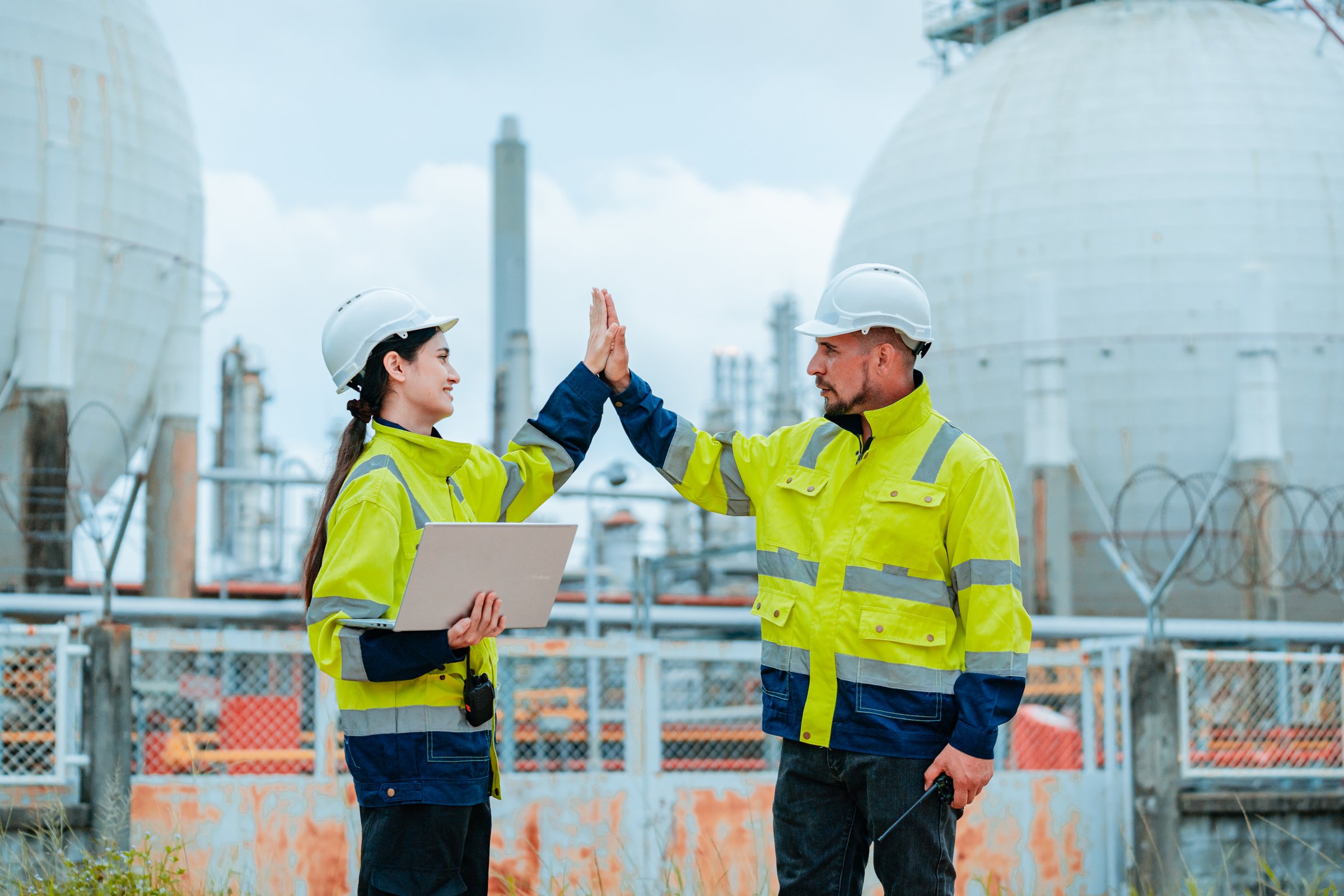 Two engineers share a moment of success on an industrial site, high-fiving each other while wearing safety gear under a cloudy sky. They hold equipment and appear proud of their work.