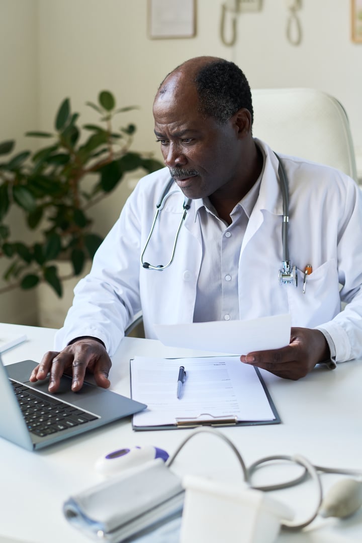 Senior doctor working in medical office, viewing patient records on laptop, reading medical chart with stethoscope draped around neck, showing serious expression