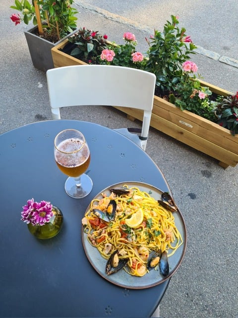 Plate of seafood pasta with lemon wedge and beer glass on a table, surrounded by decorative flowers and planters.