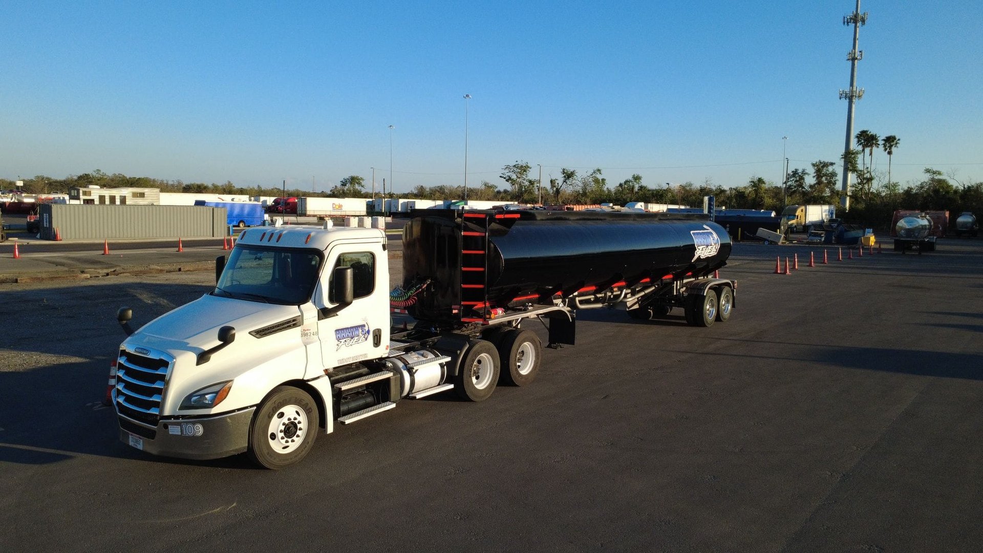 A white semi-truck with a cylindrical black tanker trailer parked on a large paved area. Traffic cones and buildings in the background.