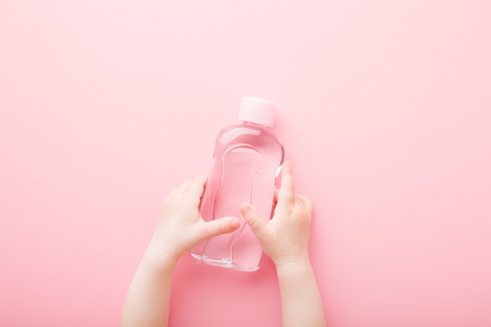 Baby girl little hands holding transparent plastic bottle of oil on light pink table background. Pastel color. Care about soft body skin. Closeup. Point of view shot. Top down view.