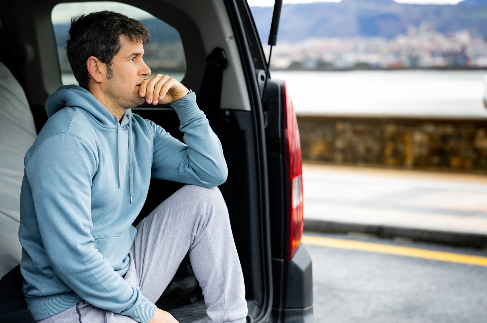 A man sits in the open trunk of a car, lost in thought as he looks out at the sea, embodying a moment of travel and reflection.