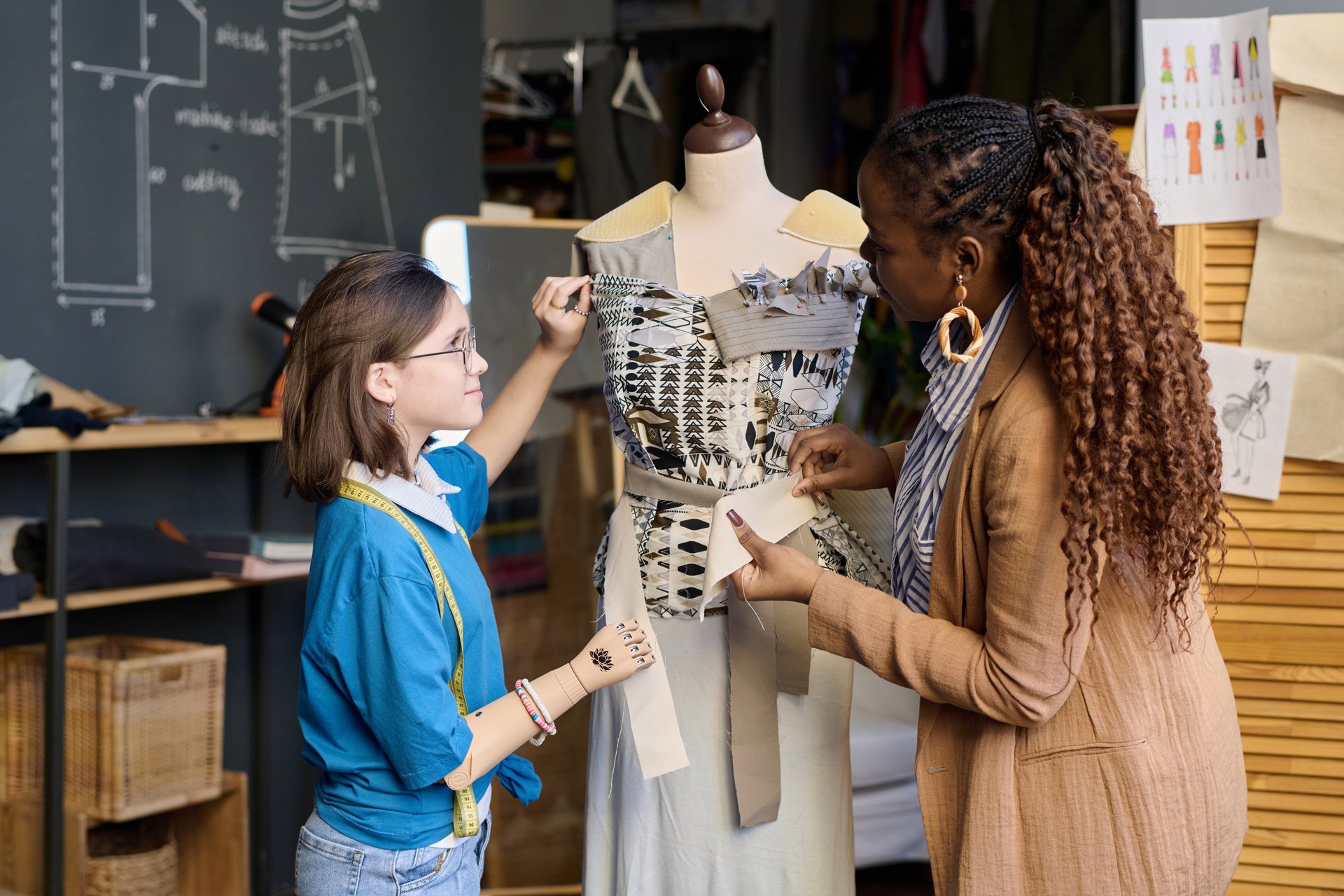 Side view portrait of female tailor teaching girl with disability clothing design and fitting mannequin together in atelier