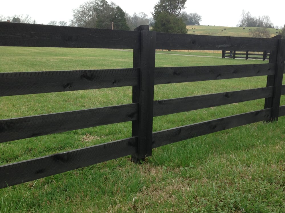 A black wooden fence with horizontal planks running across a grassy field on a cloudy day.