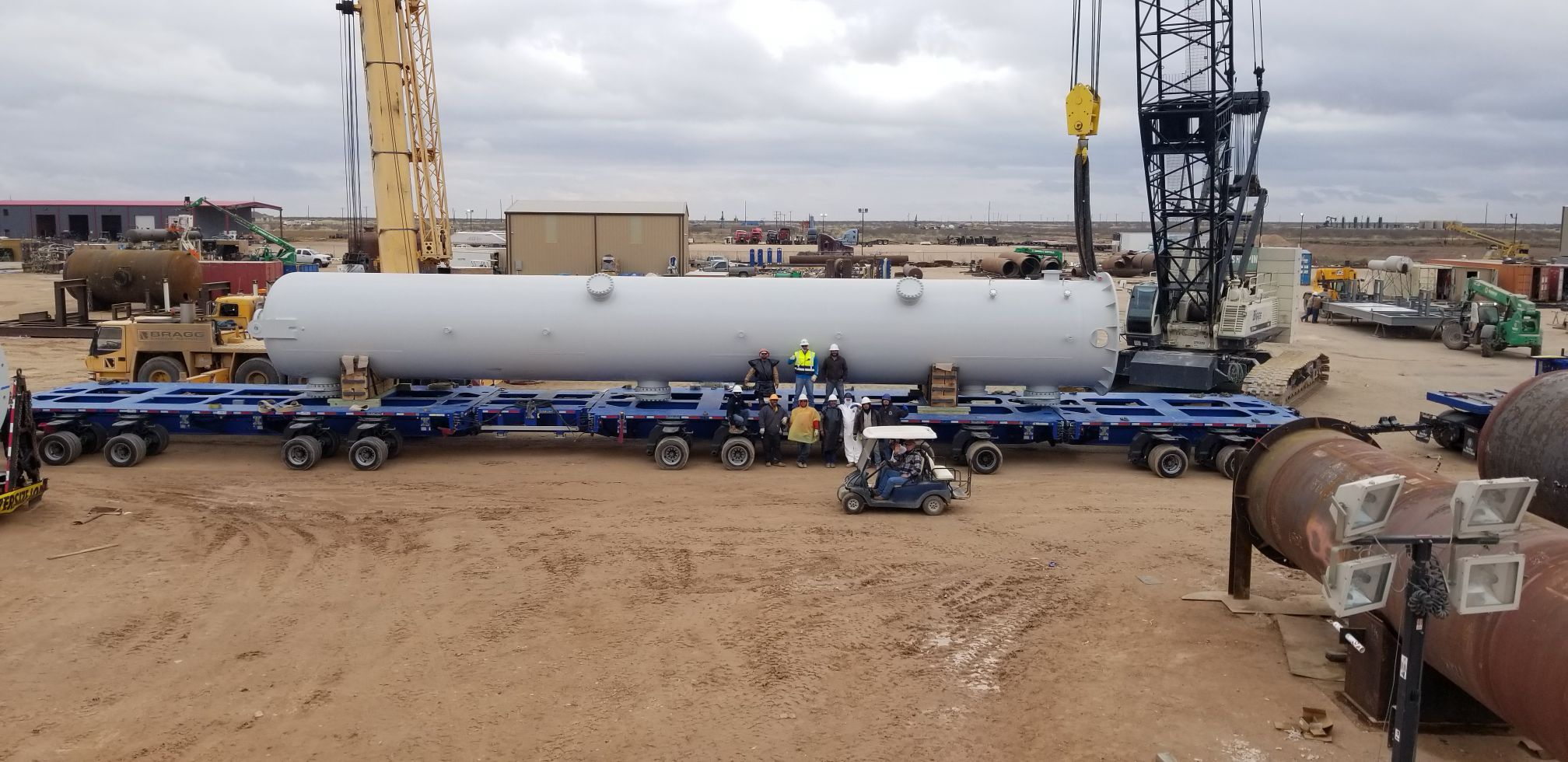 Workers and cranes handle a large industrial cylinder on a multi-wheeled platform at a construction site.