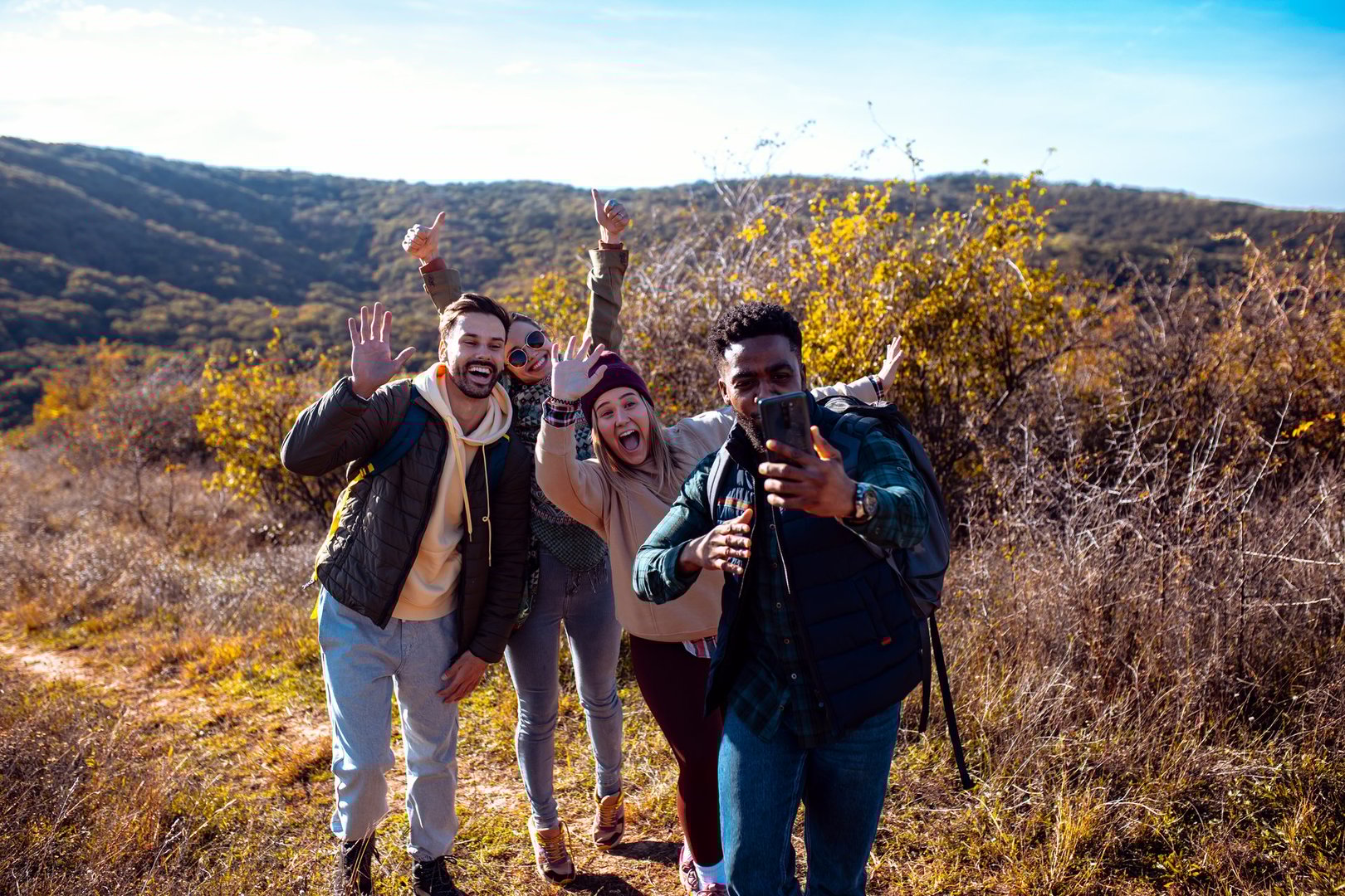 Smiling group of friends having fun while hiking together on hill at sunset and making video blog with smart phone.