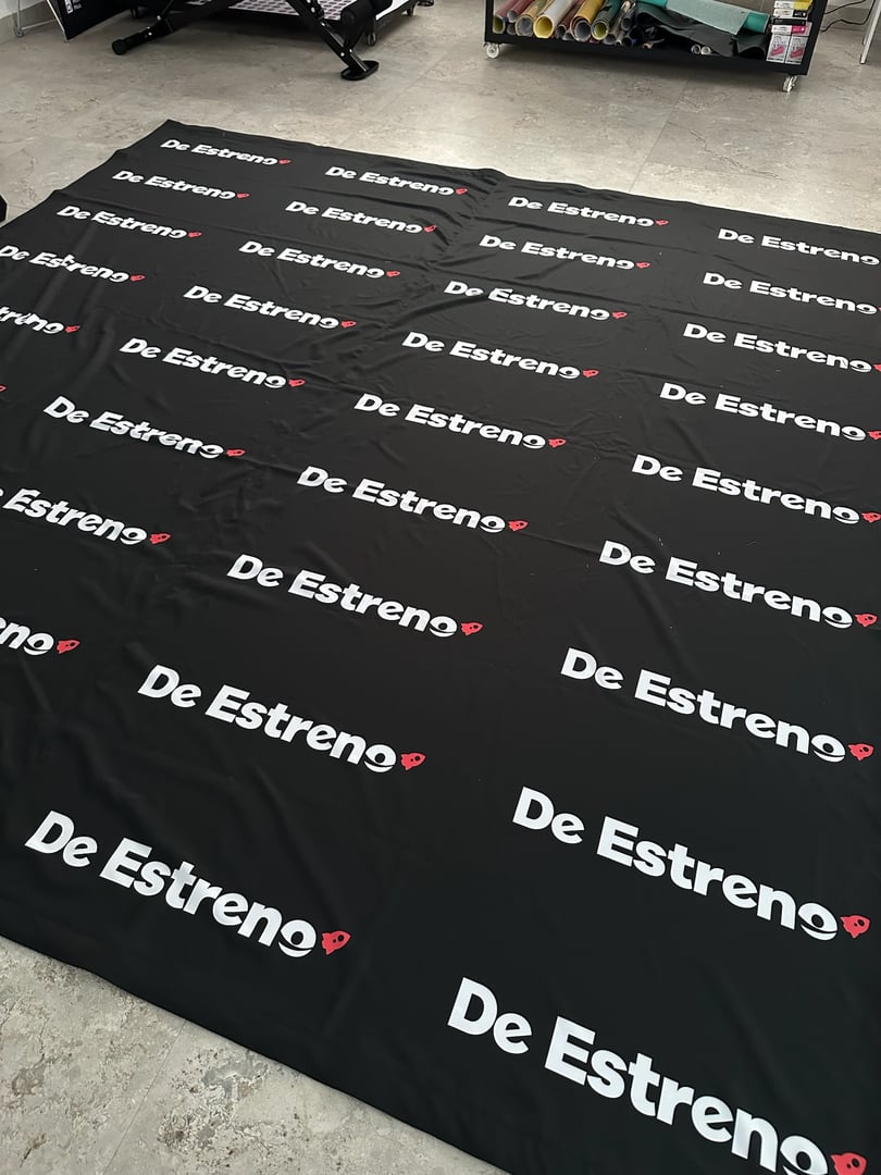 Black backdrop with repeated "De Estreno" text and a small red symbol, laid out on a light-colored floor.