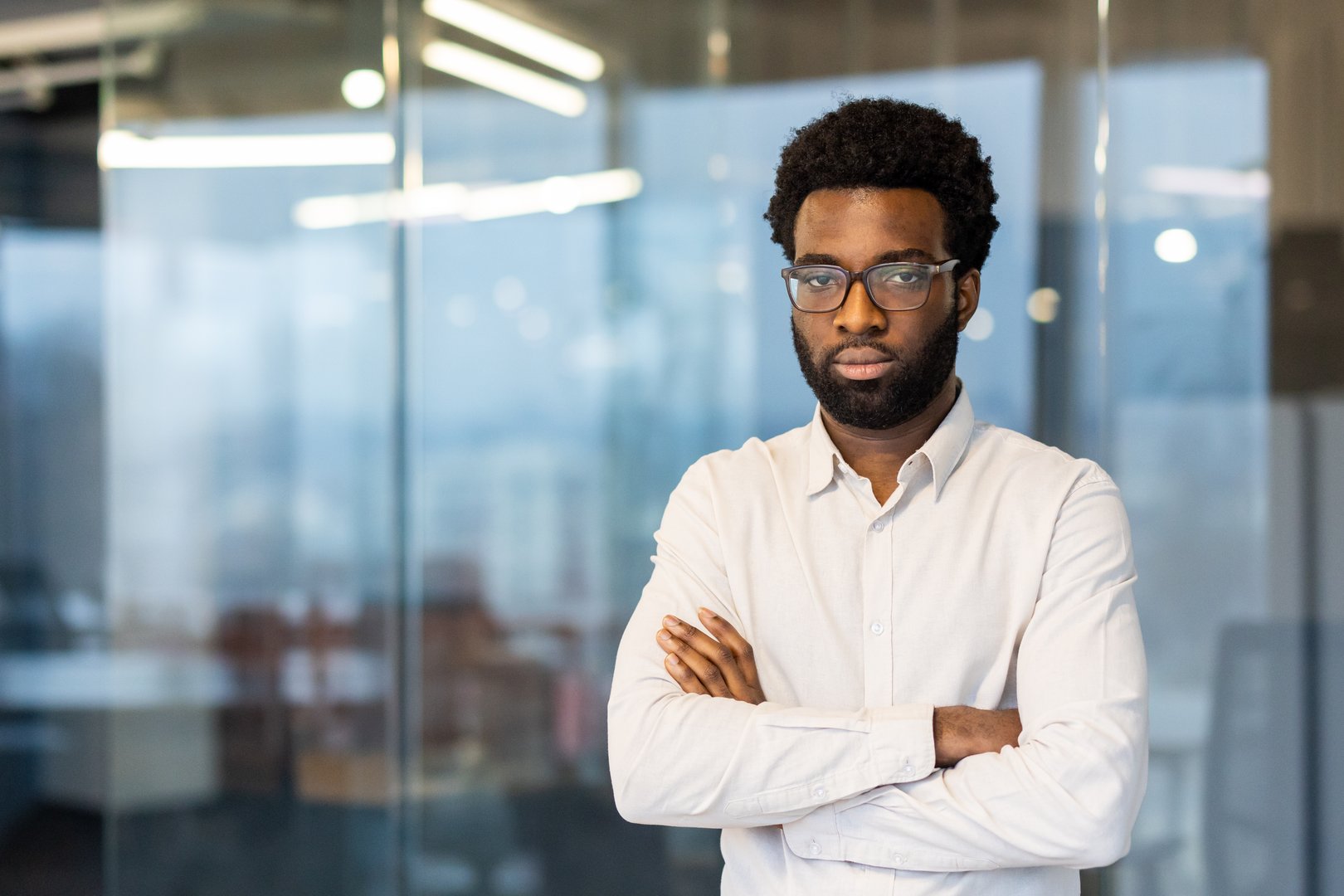 Young african american businessman wearing glasses, crossing arms confidently in a modern office setting, exuding professionalism and determination amidst a stylish urban backdrop