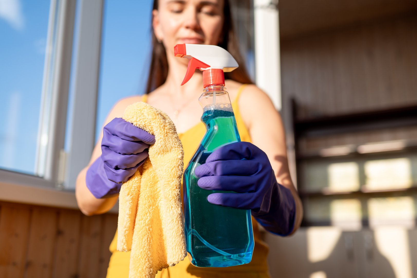 Close-up of gloved hands of a young housewife holding window cleaning spray and cloth, ready for household chores