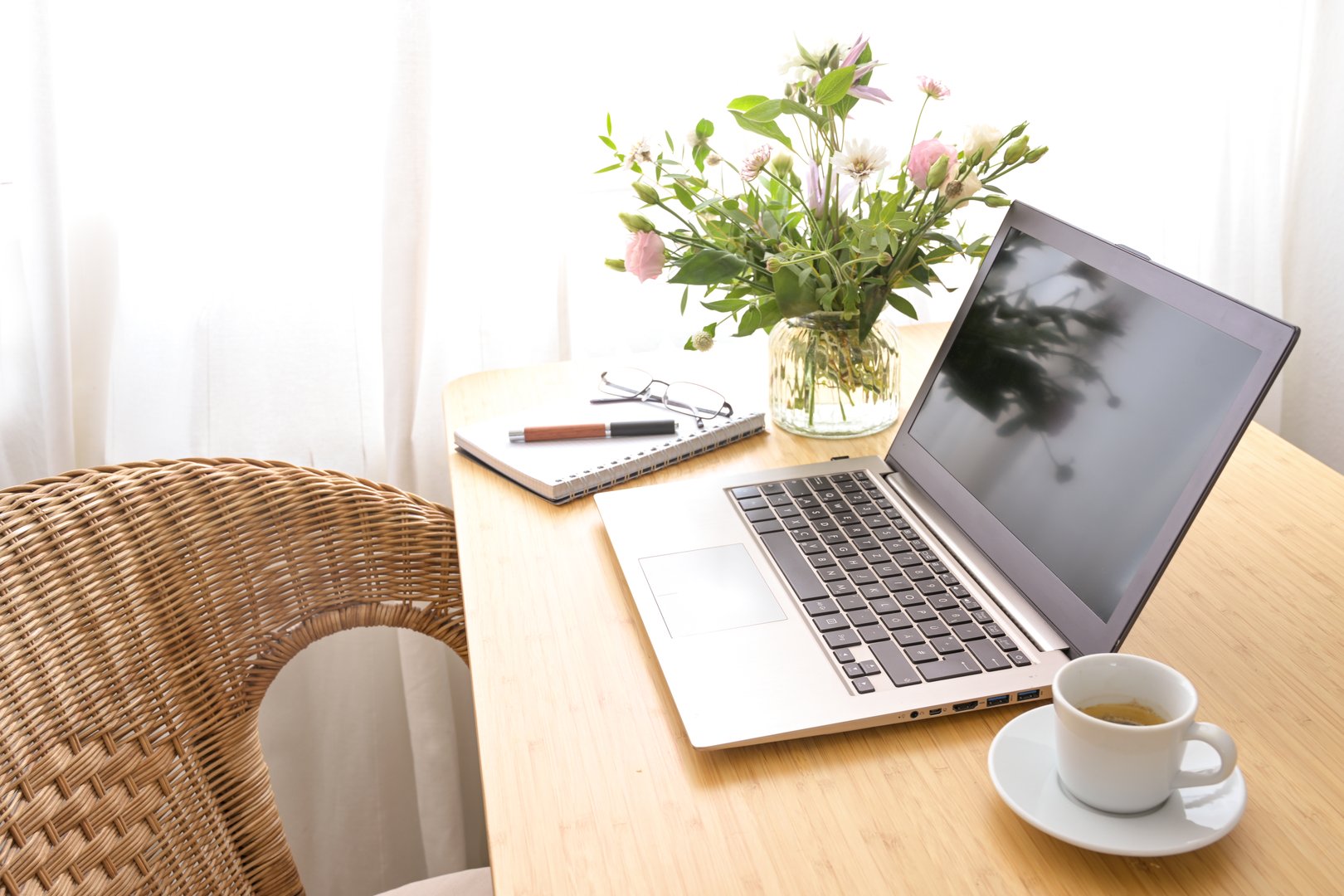 Laptop computer, coffee, flower bouquet and stationery on a wooden table with a wicker armchair, in a feminine, warm and cozy home office, creative working business, copy space, selected focus