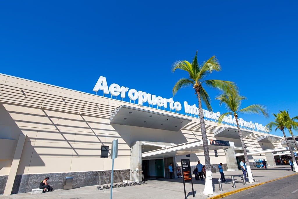 Exterior of an international airport with palm trees, featuring a sign reading Aeropuerto Internacional Puerto Vallarta.