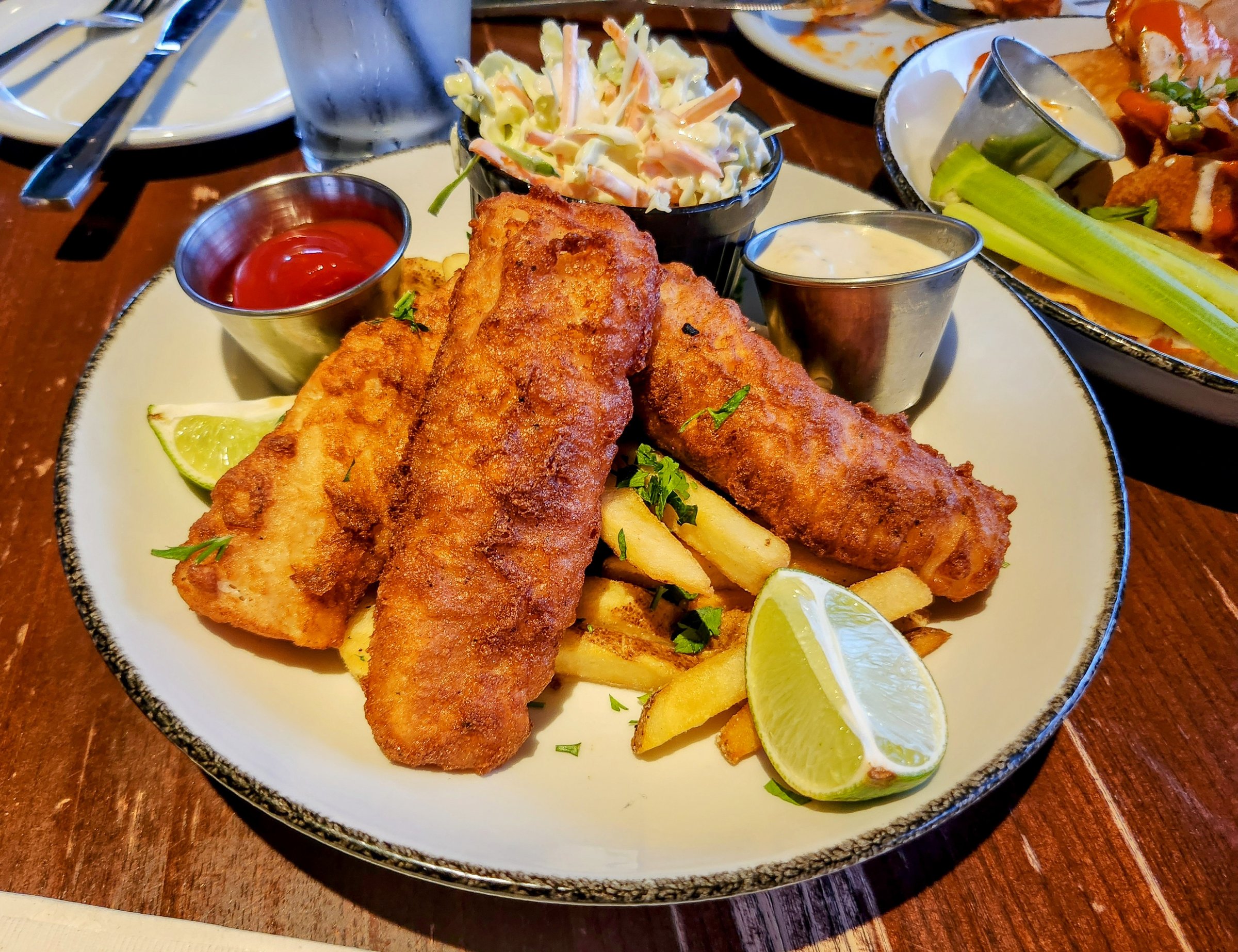 Three pieces of golden fried cod fish sitting on a bed of french fries and served with coleslaw.