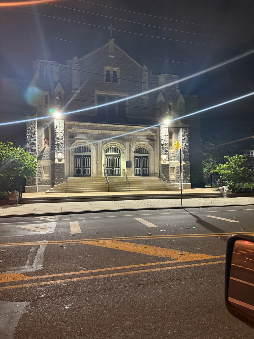 Historic stone church facade illuminated at night with three large doors, ornate architecture, and dramatic spotlight beams crossing the front.