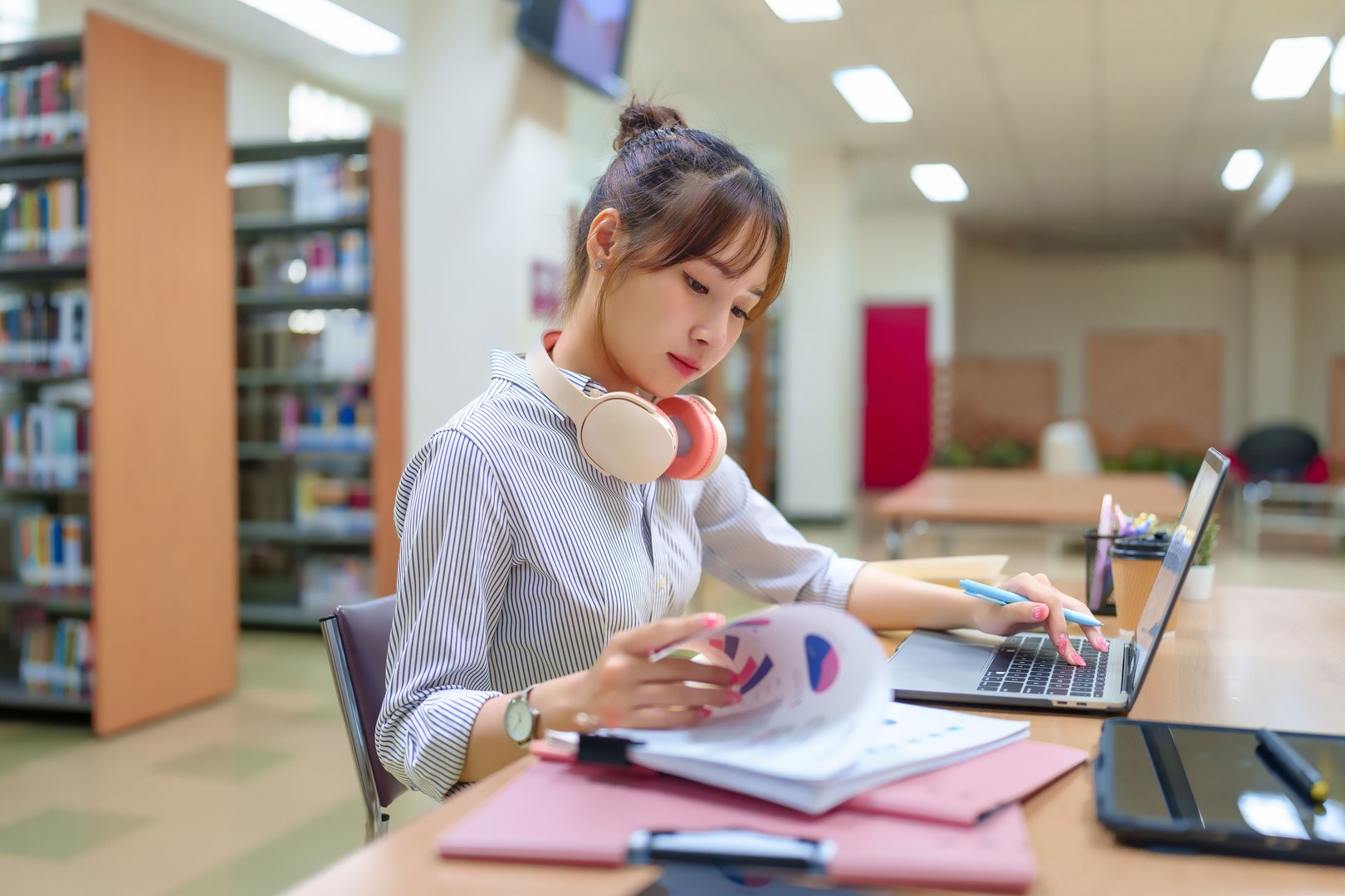 A young Asian woman is focused on studying with her laptop and analyzing charts in a library. She is wearing headphones and surrounded by books and educational materials, showcasing her dedication to learning and research.