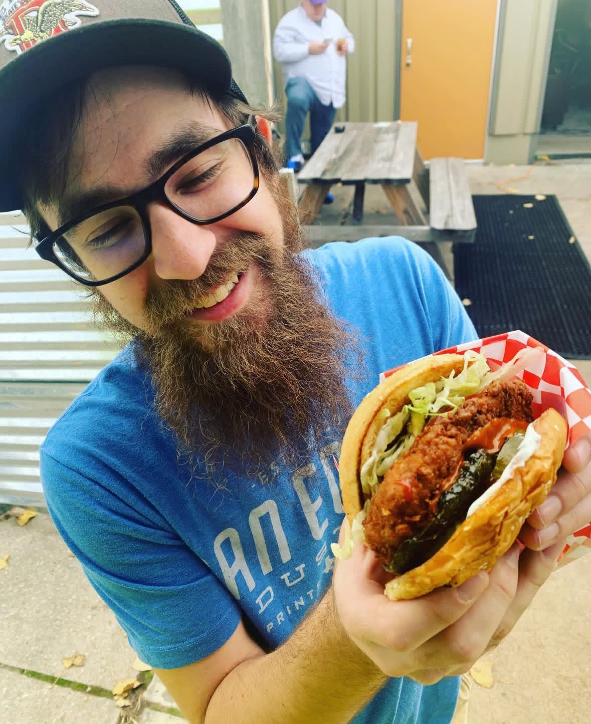 Man with glasses and beard smiling while holding a large burger with lettuce and sauce.