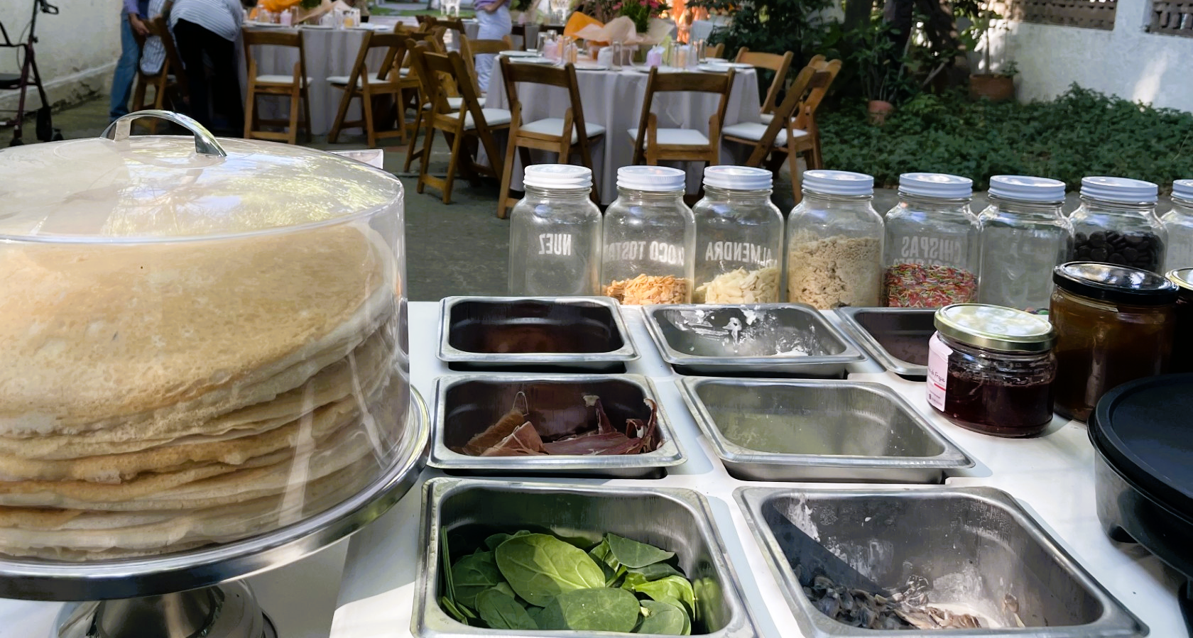 Pancake station with toppings and condiments; tables set for an outdoor event in the background.
