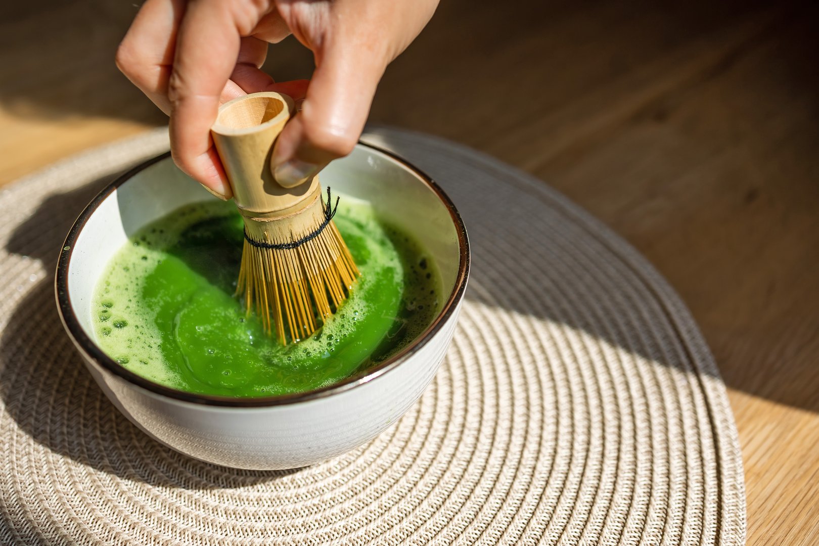 A hand using a bamboo whisk to froth vibrant green matcha tea, representing Japanese culture and tradition. The tea is prepared in a ceramic bowl, highlighting the art of tea-making.