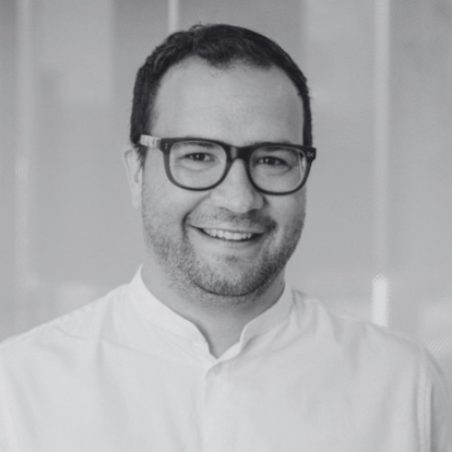 Smiling man with glasses and a beard wearing a light-colored shirt, photographed in black and white.