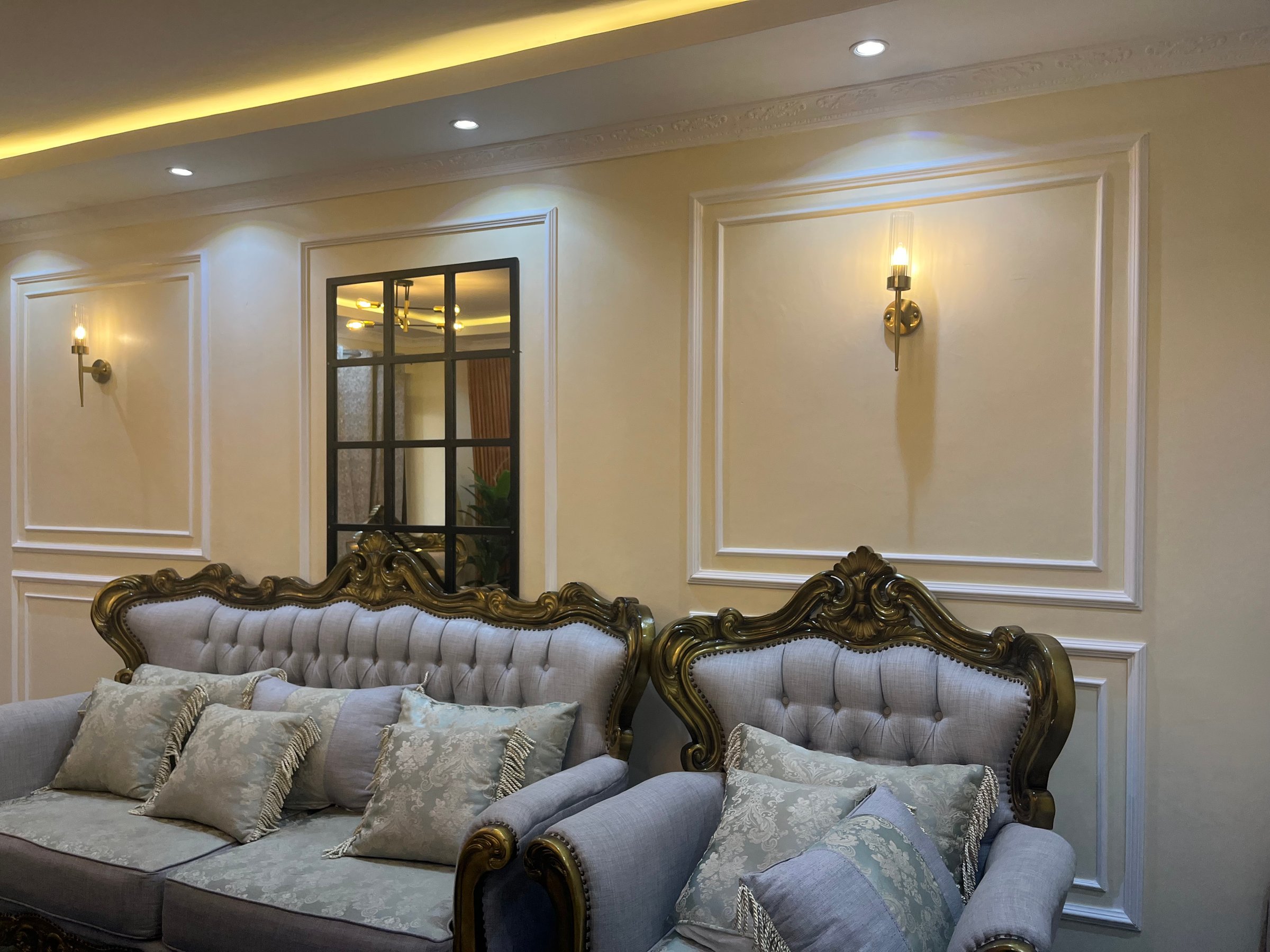 Elegant living room with ornate armchairs, a large mirror, sconces on cream walls, and a plush grey sofa with patterned pillows.