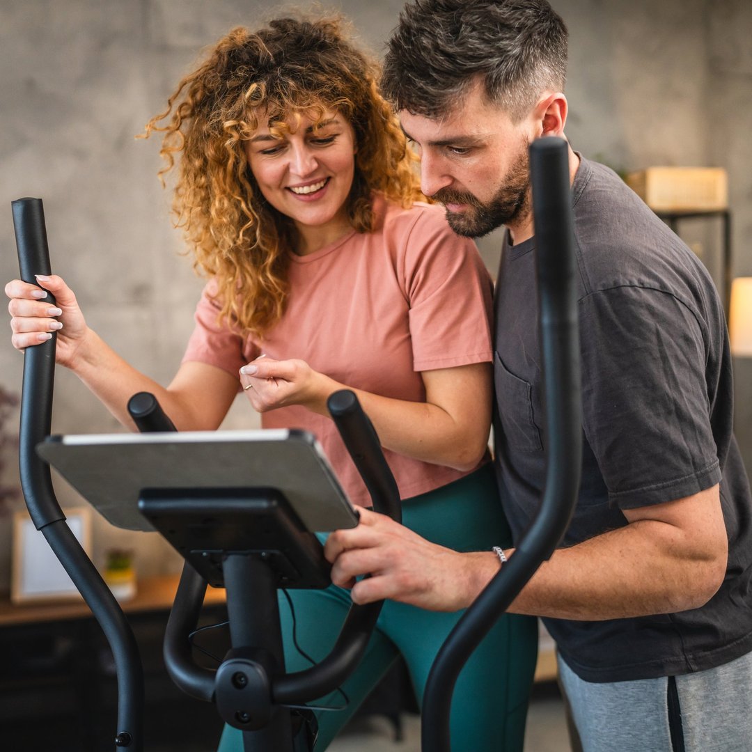 woman exercise on elliptical machine and follow training on tablet, while her boyfriend help her