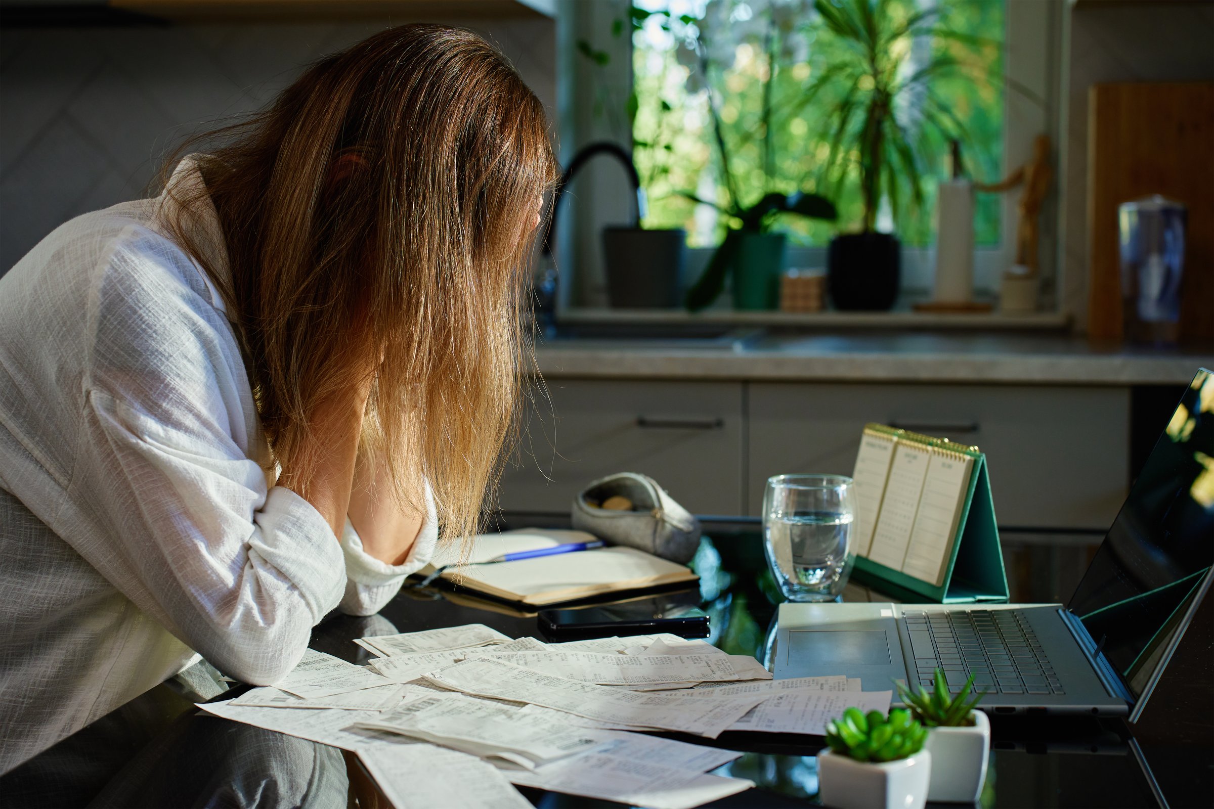 Stressed woman reviewing bills at home. Female organizing receipts, sitting at kitchen table with laptop and paper documents. Concept of budgeting and managing personal finances