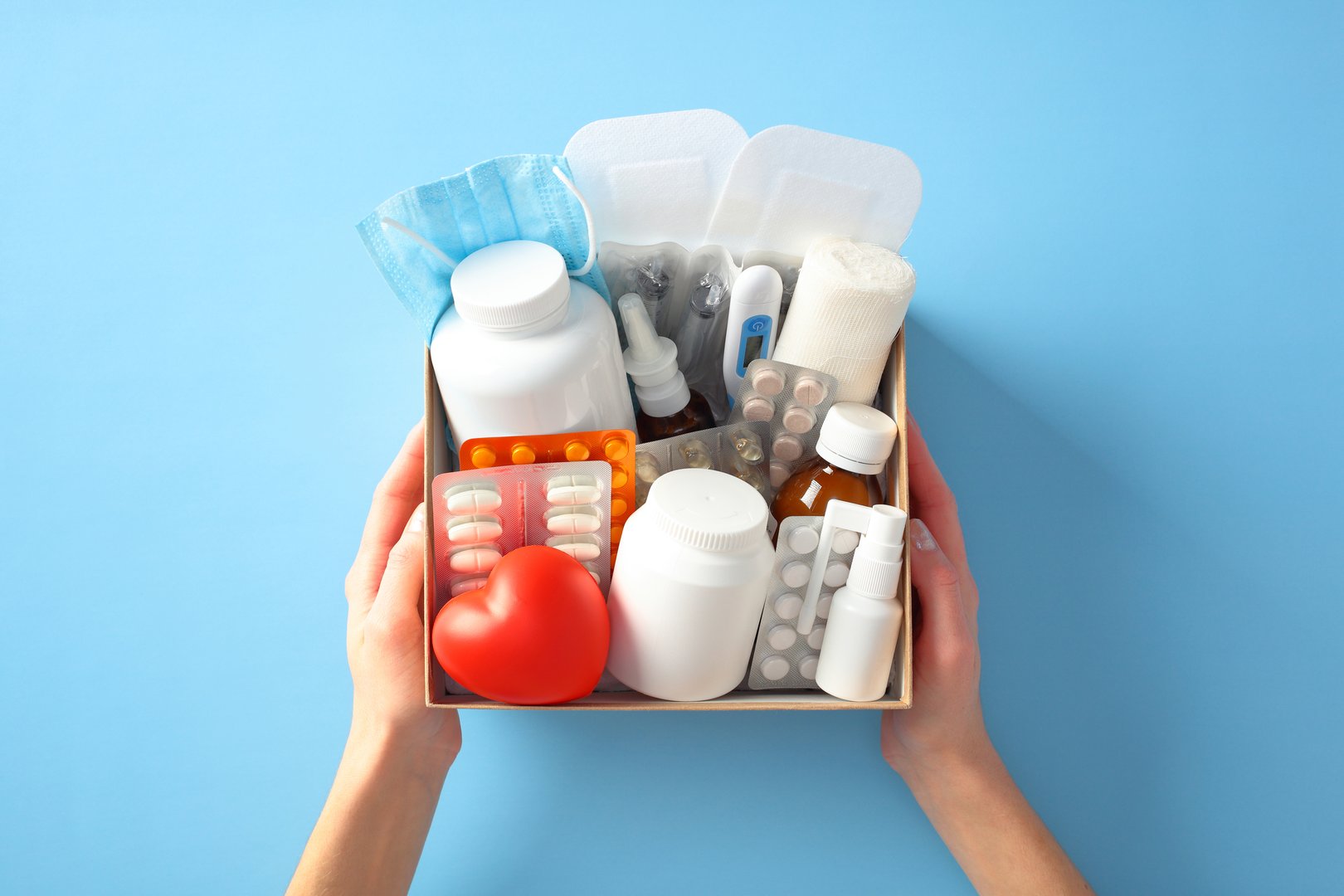 Female hands holding medical supplies box with various items including pills, bottles, red heart, a thermometer, and a face mask on a blue background. Healthcare and emergency preparedness concept