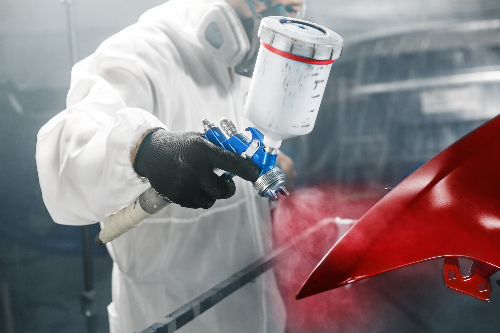 Car painter sprays red color paint to part of vehicle using spray gun in workshop garage. Man working in paint chamber. Complex repair and restoration automobile with exact select of color after accident.