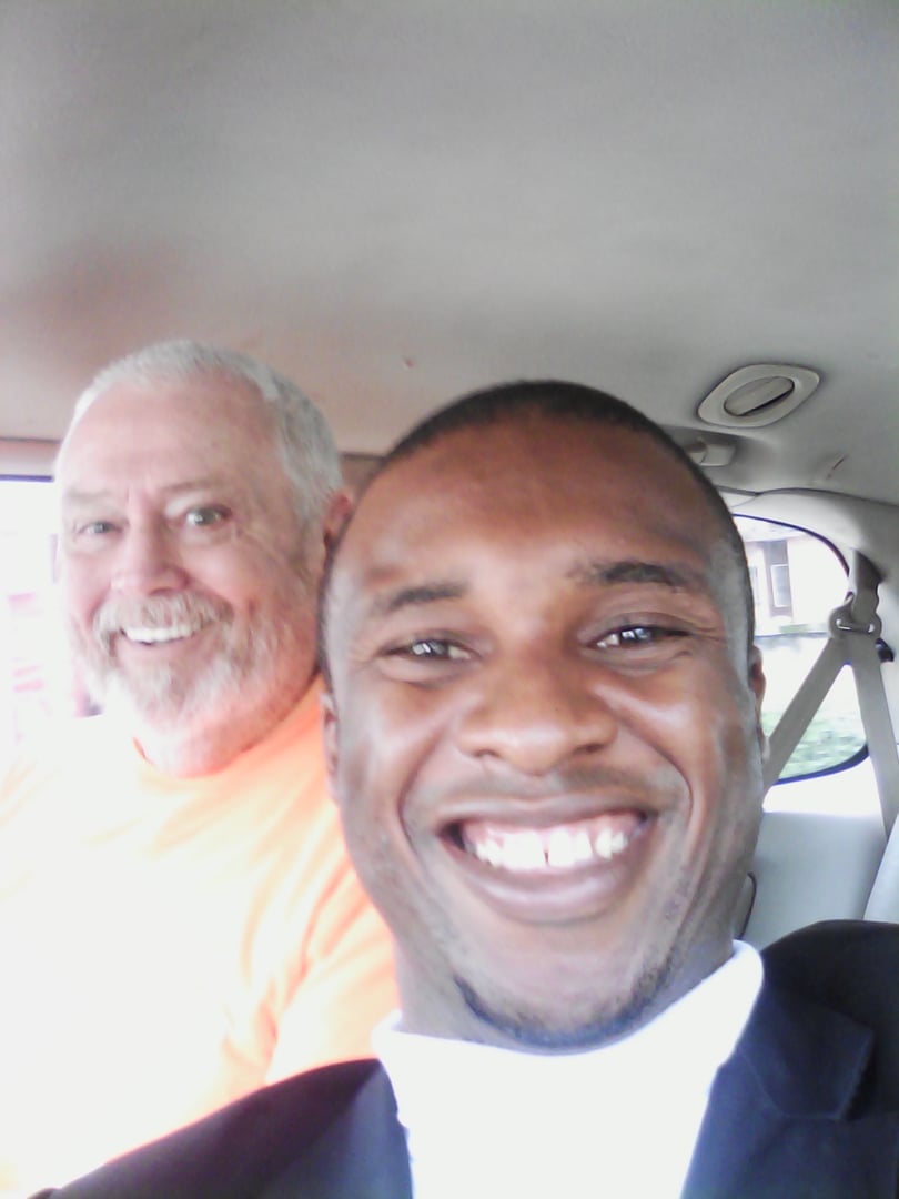 Two smiling men taking a selfie in a car, one wearing an orange shirt and the other in a dark jacket with a white shirt.