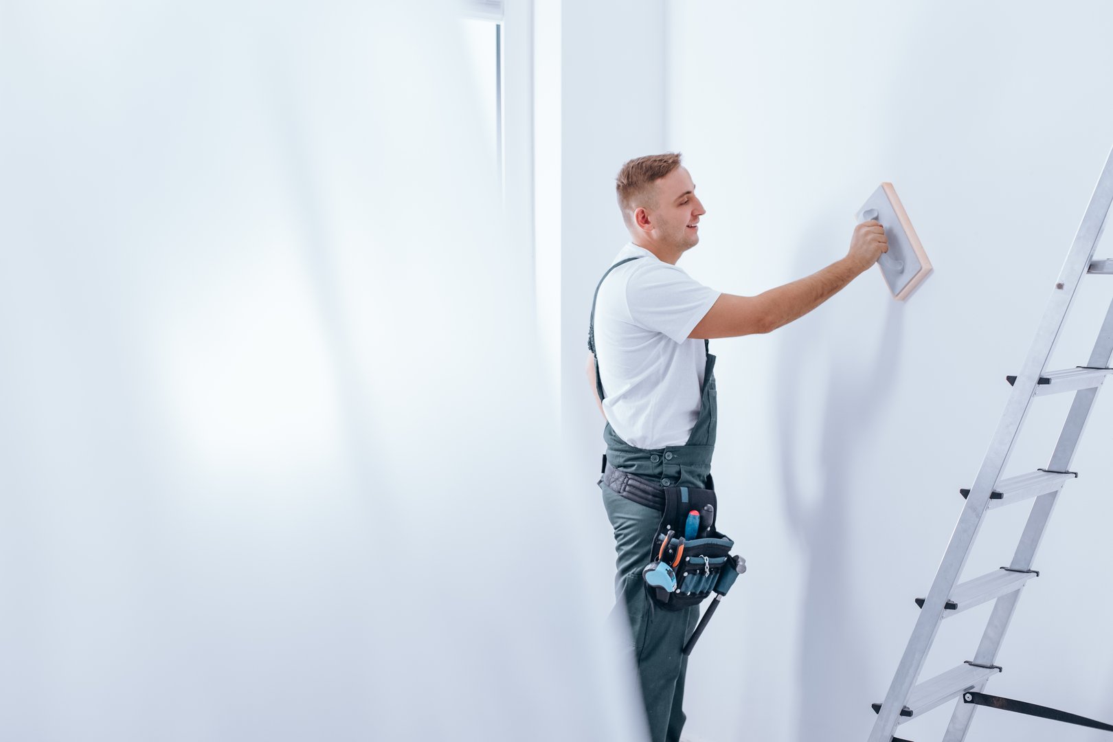 Young smiling painter with tool belt pinned to his workwear smoothing the wall before painting