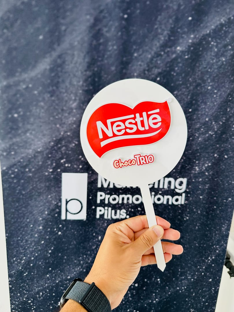 Person holding a Nestlé Choco Trio sign in front of a background with text indicating marketing or promotion.