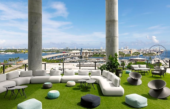 Rooftop lounge with modern furniture, overlooking a cityscape and waterfront, featuring large concrete pillars and a Ferris wheel.