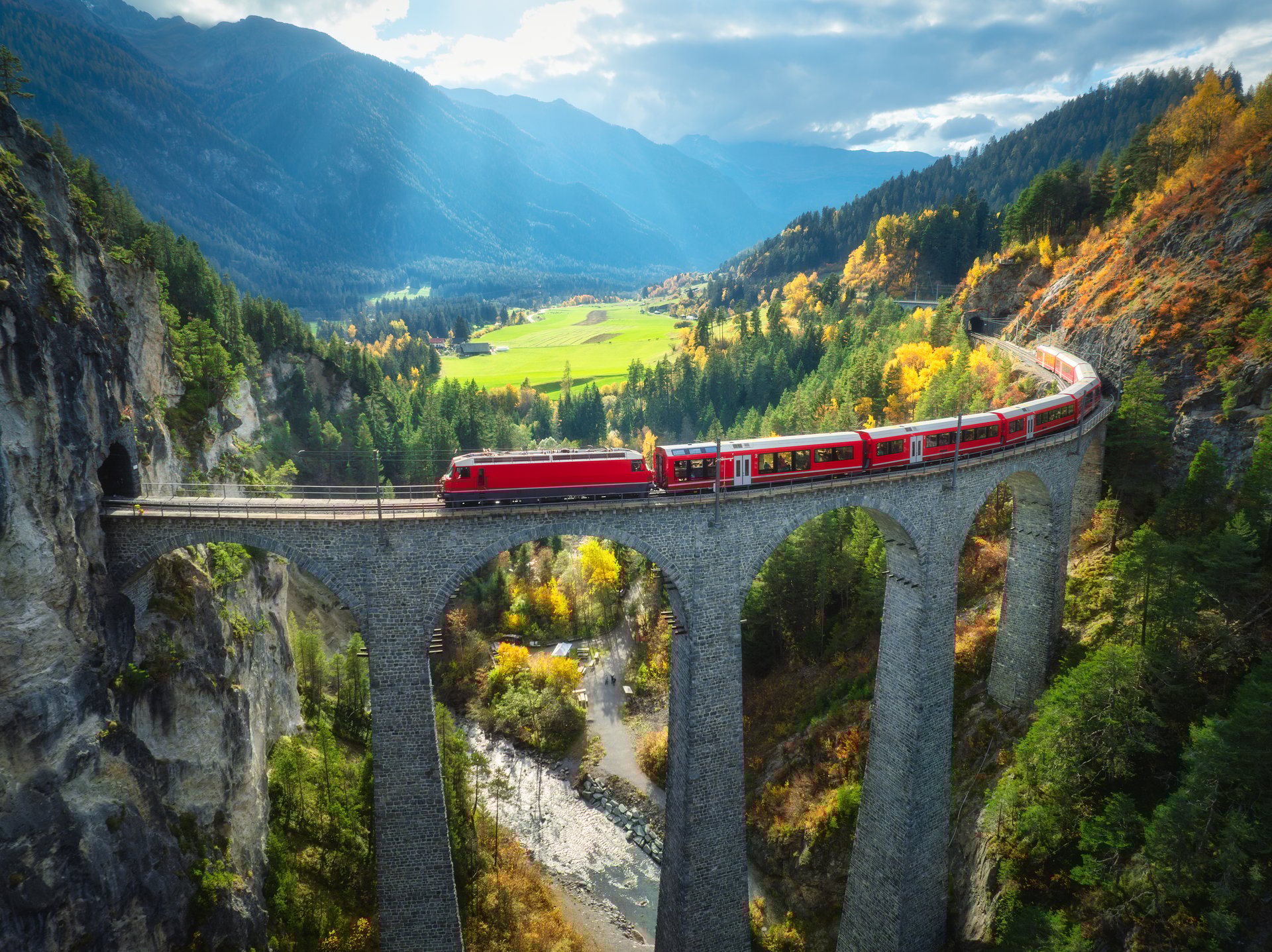 Aerial view of modern red train on Landwasser viaduct in alpine mountains, colorful forest at sunset in autumn. Bernina Express, Switzerland. Top view of train in Alps, railroad, green trees in fall