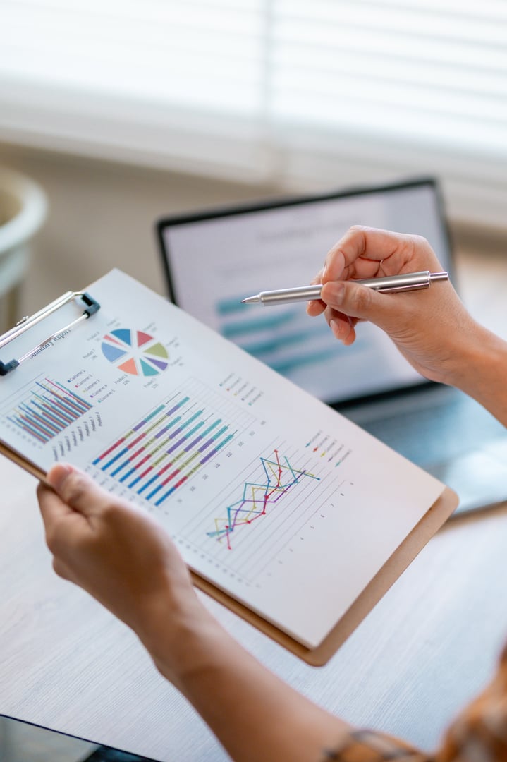 Businesswoman pointing at colorful charts and graphs on a clipboard while comparing them with data displayed on a laptop, showcasing financial analysis and business strategy development