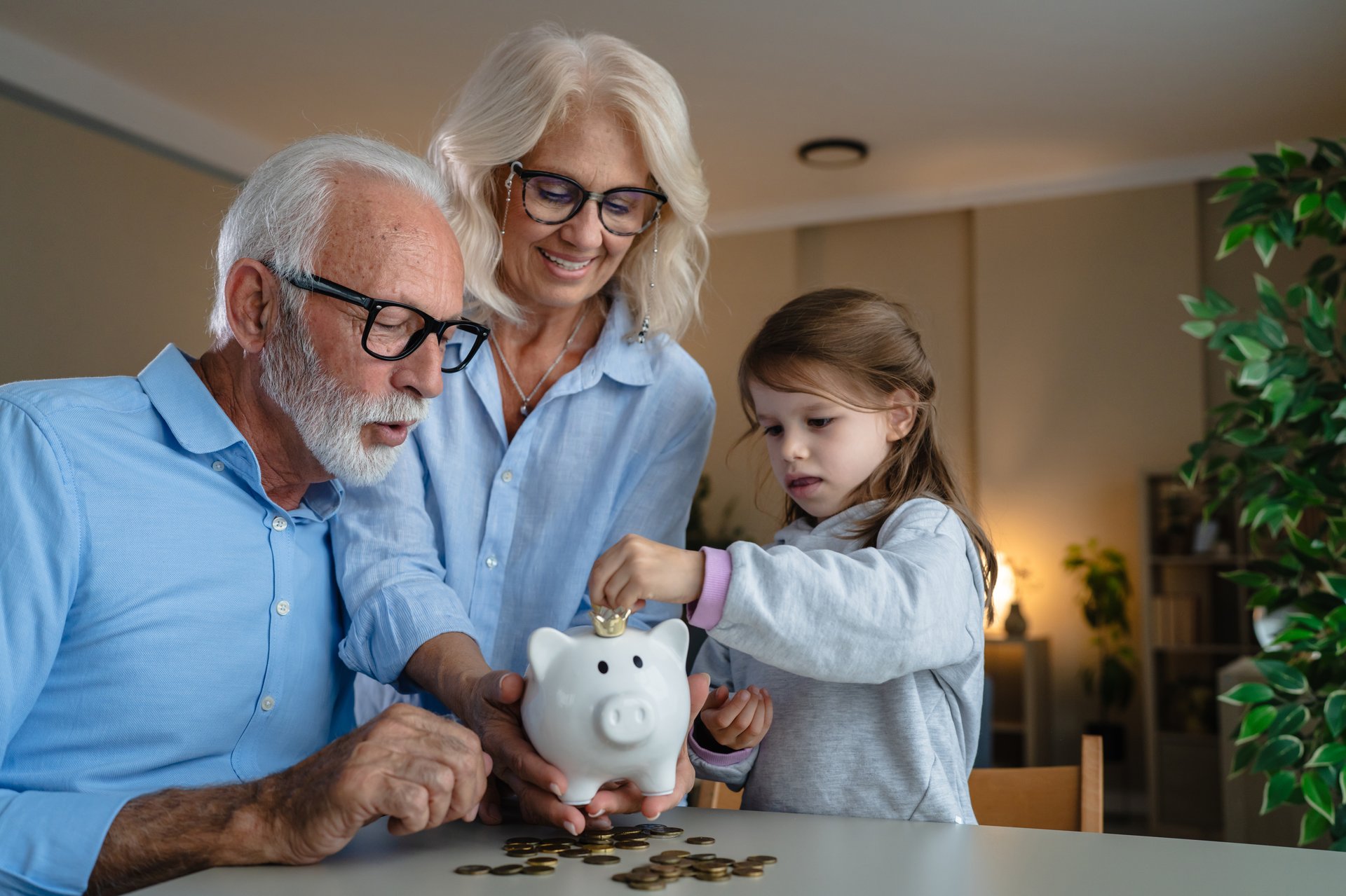 Grandparents are teaching their young granddaughter about financial literacy and the importance of saving money by putting coins into a ceramic piggy bank together at home
