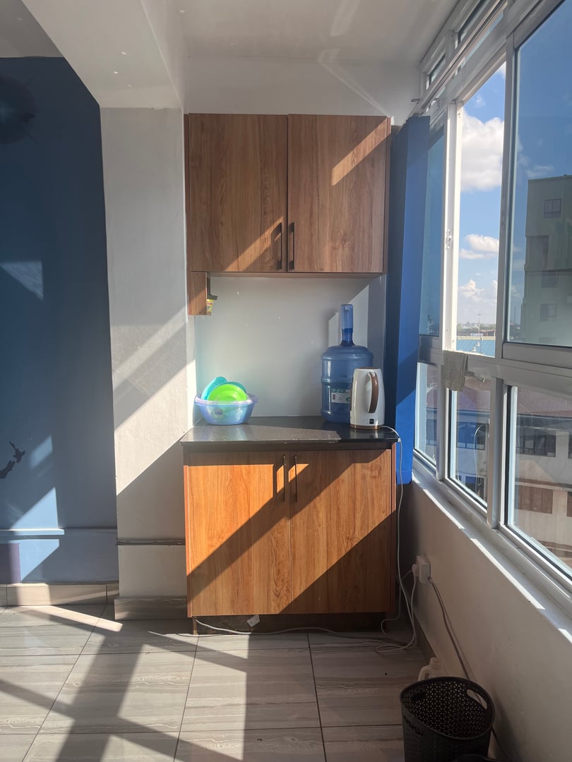 Kitchen corner with wood cabinets, water dispenser, electric kettle, and a bowl of plastic dishes near a window.