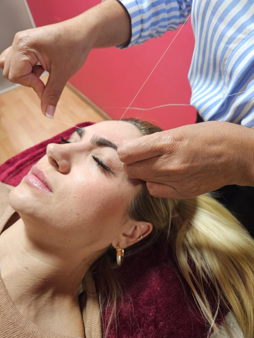 Person receiving eyebrow threading in a salon, lying on a red towel. Thread held by another person with striped sleeves.
