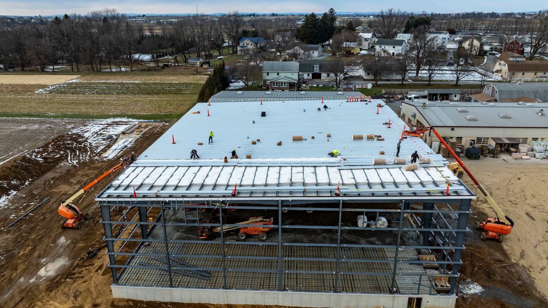 Workers are busy installing a roof on a large commercial building under construction in a rural area. The surrounding landscape shows houses and fields, with snow visible on the ground.