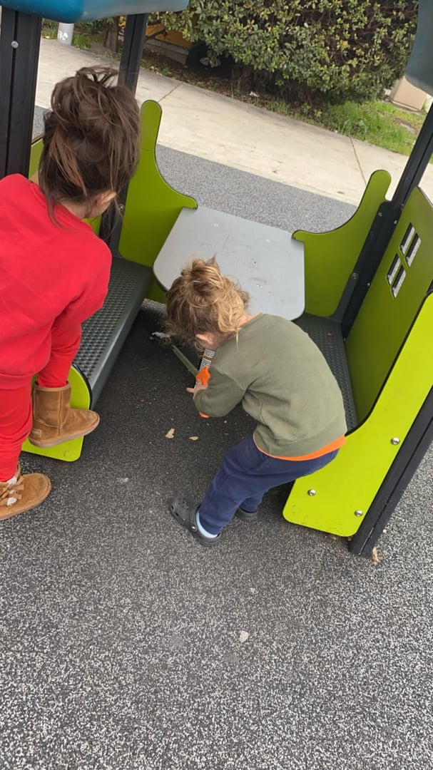 Two children play on a playground structure. One child in red looks on while the other in green bends down to explore.