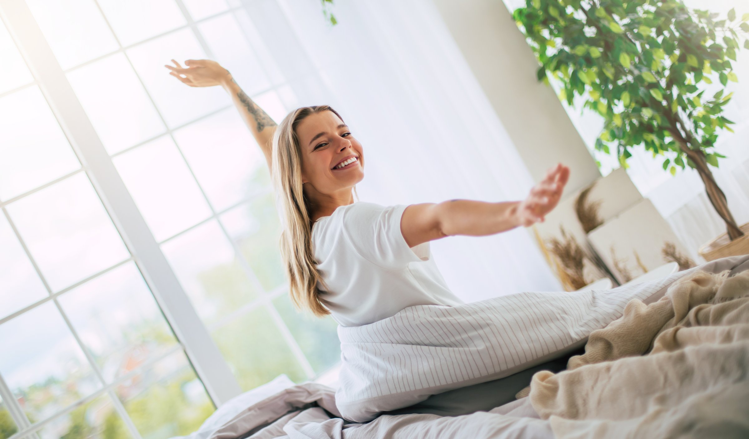 Cute smiling woman stretches joyfully on a bed, bathed in natural light from large windows. The bedroom is bright and cozy, with plants and soft bedding, evoking a sense of comfort and happiness