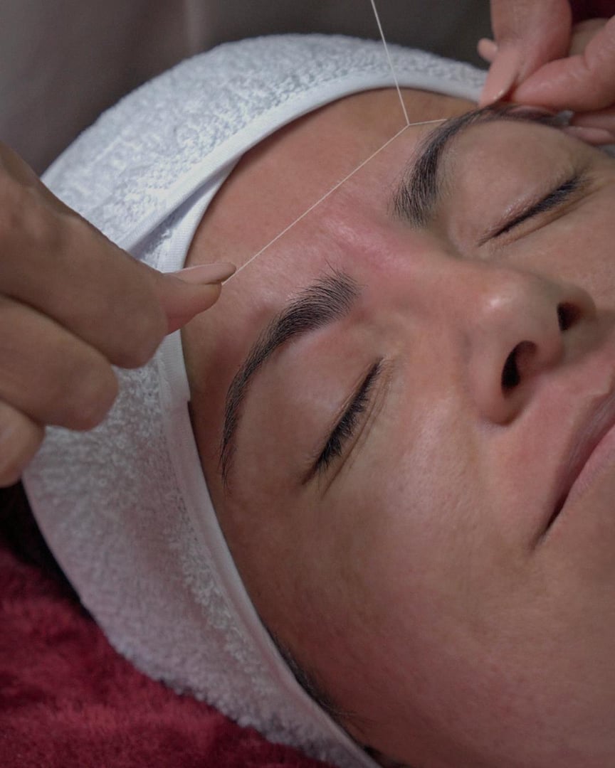 Person receiving eyebrow threading, lying with eyes closed, wearing a headband, as hands hold the thread.