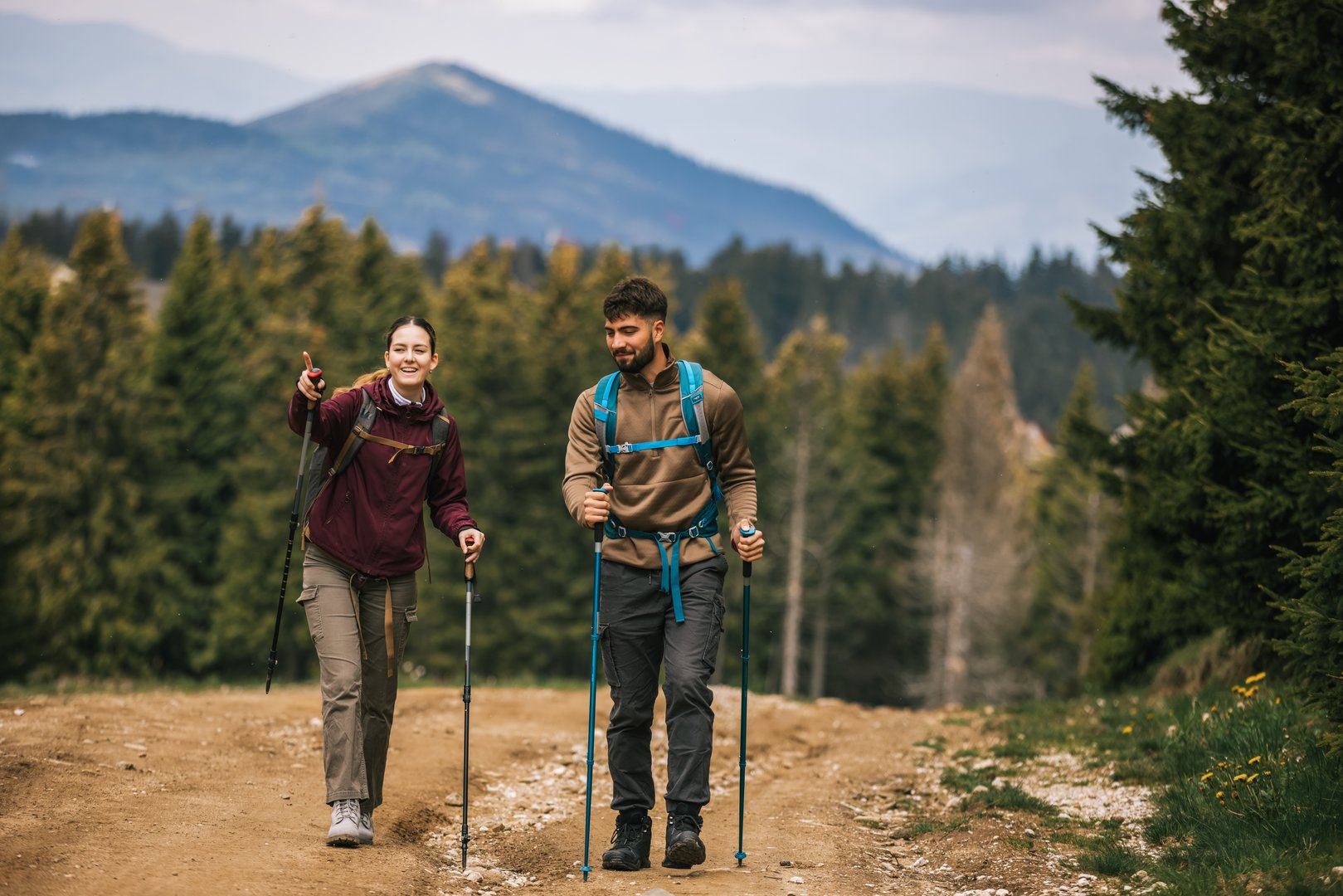 A young man and woman are hiking through a scenic mountain forest, fully equipped with backpacks, hiking poles, and proper outdoor clothing. They pause on the trail to look at something in the distance, sharing a moment of curiosity and discovery.