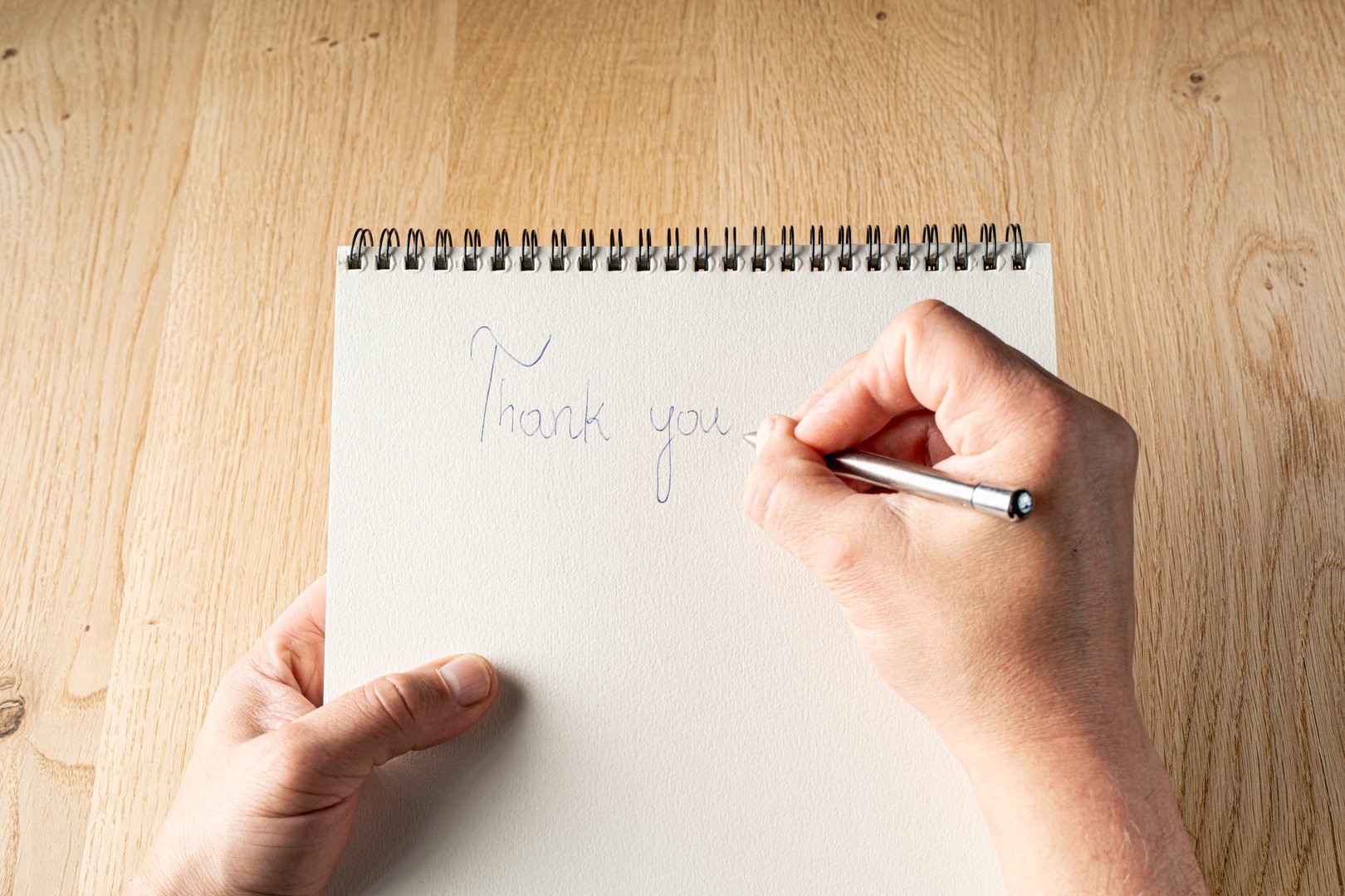 A person is writing a thank you note on a spiral bound notebook. The person is using a pen to write the note