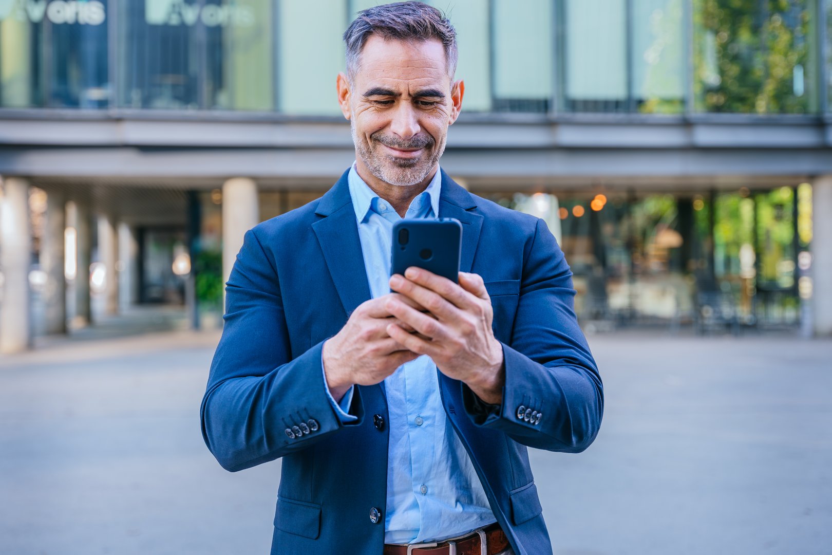 Smiling middle-aged businessman in a blue shirt holding and using a smartphone outdoors in a modern urban setting.