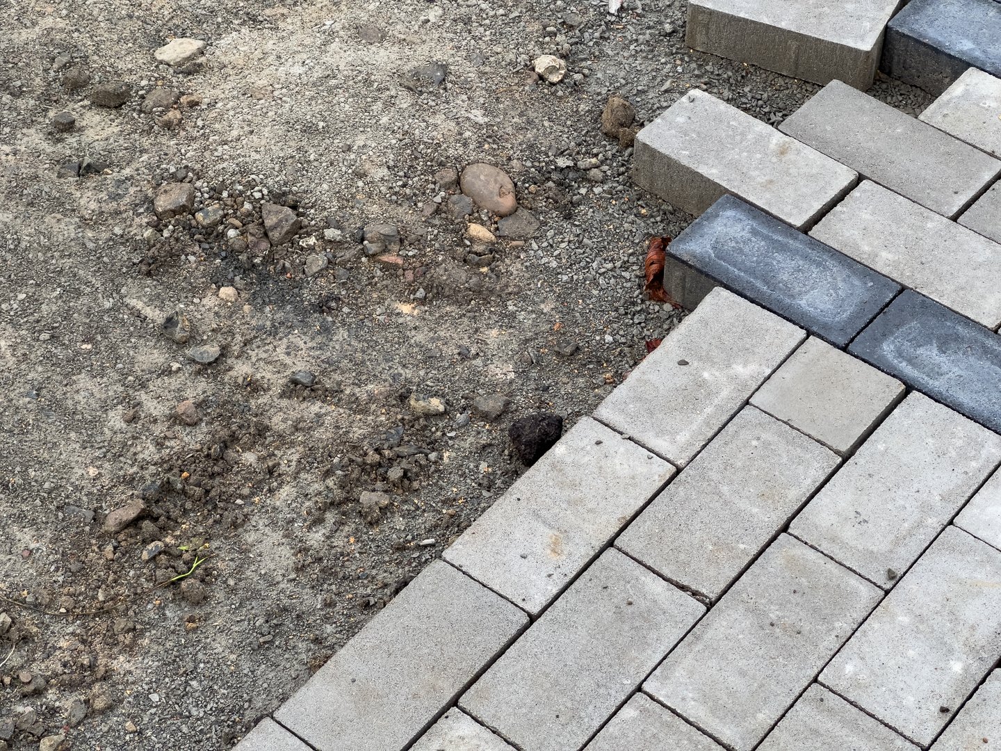 A section of pavement is being constructed, showcasing gray interlocking bricks. Nearby, an unfinished area of gravel and loose stones is visible, indicating progress in development.