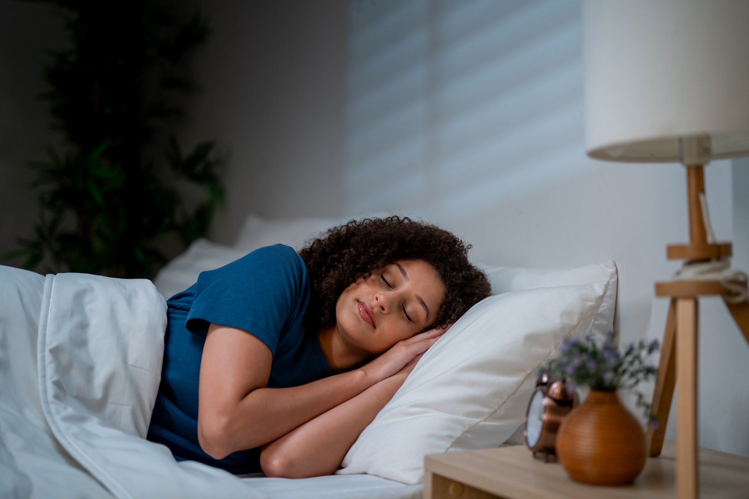 Attractive Latina woman lying on bed in a dark bedroom at night. Young female feeling happy and relaxed, resting with cozy pillow and blanket, enjoying a peaceful holiday weekend at home in cozy room.