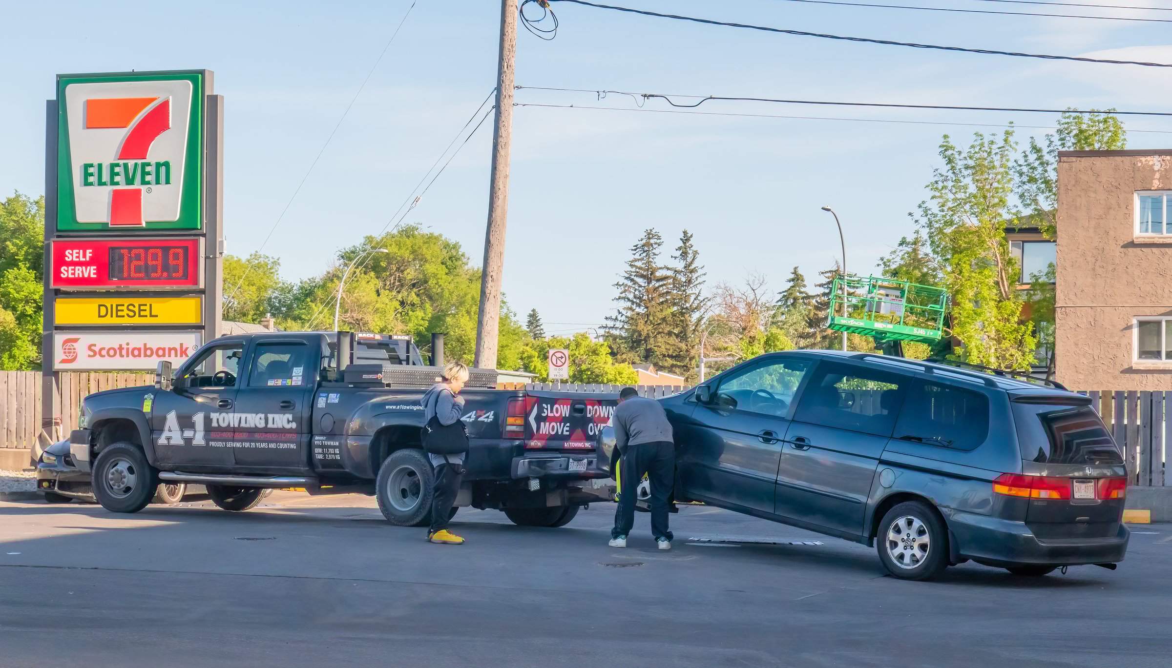 Calgary, Alberta, Canada. Jun 5, 2025. A Towing truck lifts a dark minivan at a 7-Eleven gas station, illustrating roadside assistance and essential services.