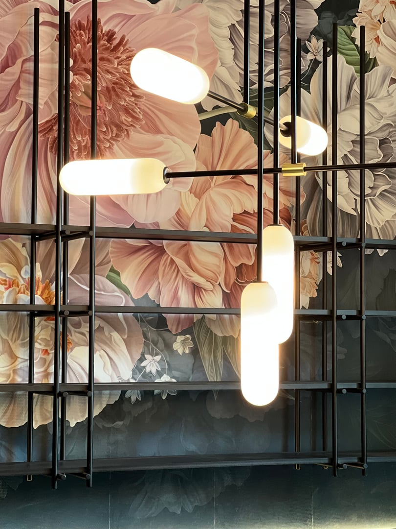 Modern hanging light fixture with tubular bulbs against a floral wall mural featuring large pink and white flowers.