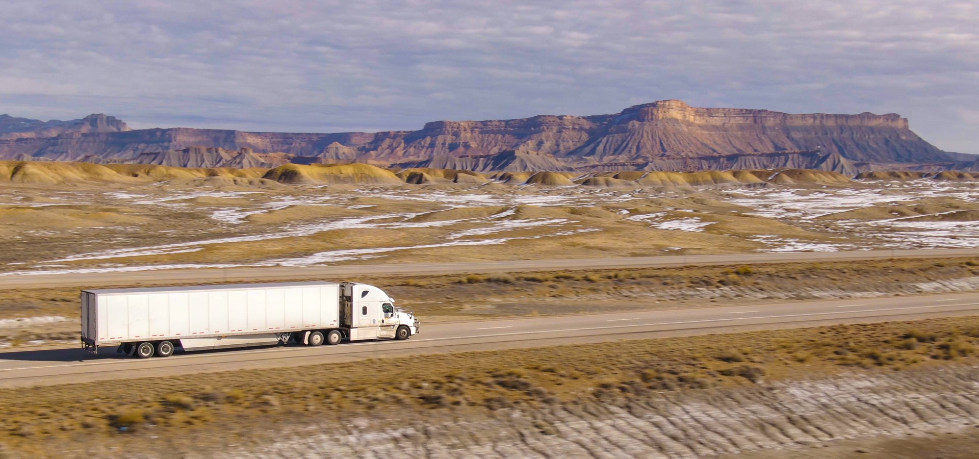 AERIAL: Semi-trailer truck speeds along the motorway running across the desert in Utah. Wintry desert landscape surrounds truck driving down the interstate highway running across rugged US wilderness.